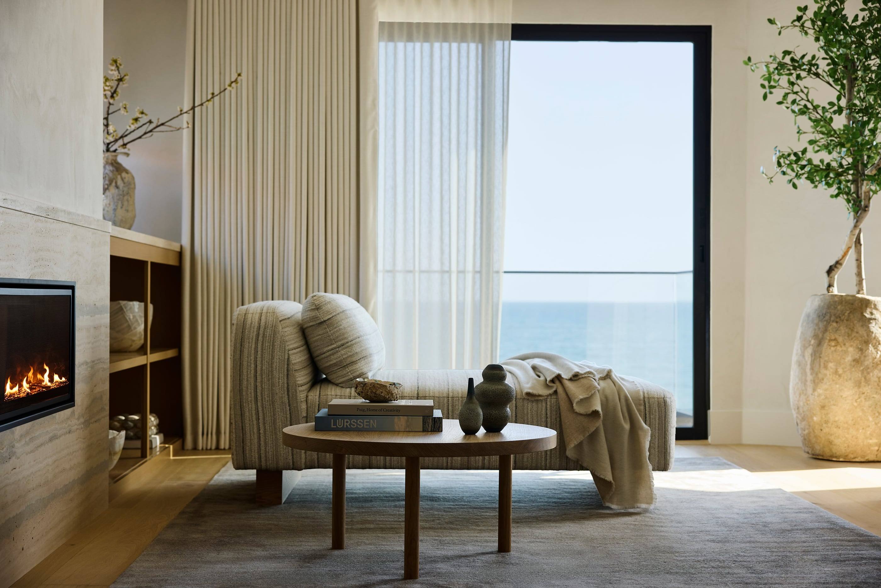 Striped chaise and round oak table before a wall of glass open to the Pacific